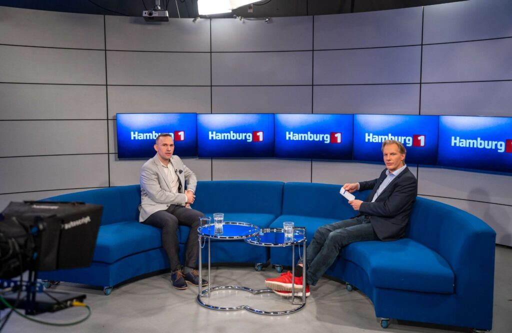 Martin Trappe sitzt auf einem blauen Sofa im TV-Studio von Hamburg 1, zusammen mit einem Journalisten. Er spricht über die wichtige Rolle von IT-Dienstleistern und die IT-Sicherheit in der heutigen Zeit.